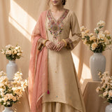 A beige women's salwar kameez suit displayed on a mannequin, featuring a pink dupatta, heavily embroidered multicolor neckline, and floral border detailing on the hem and cuffs, set against a warm beige draped backdrop with decorative flowers.