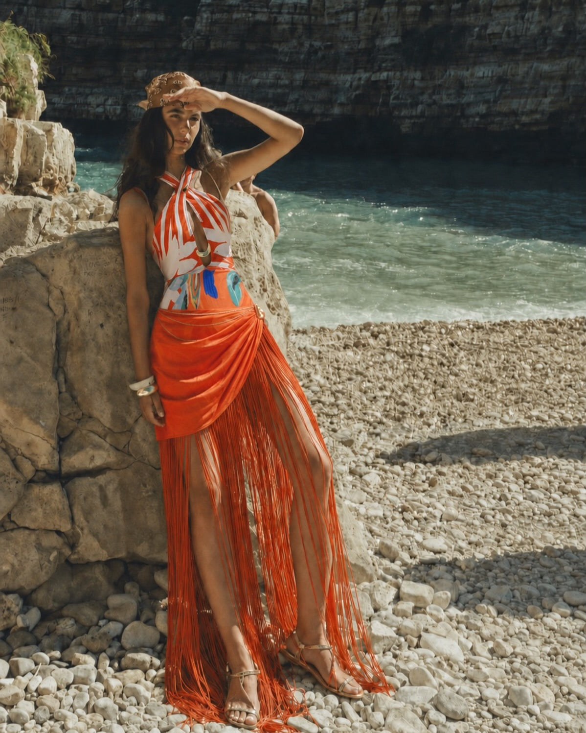 A woman stands on a rocky beach wearing a sleeveless halter dress with a white, orange, and blue abstract print and a long orange fringe skirt. The background shows water and dark cliffs. Resort Wear 