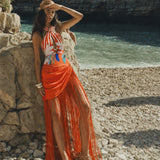 A woman stands on a rocky beach wearing a sleeveless halter dress with a white, orange, and blue abstract print and a long orange fringe skirt. The background shows water and dark cliffs. Resort Wear 