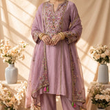 A lavender purple ethnic suit set displayed on a mannequin, featuring a kurta with colorful floral embroidery, matching trousers, and a sheer dupatta draped over a decorative stand in a warm, styled studio setting.