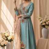 A light blue ethnic salwar kameez suit displayed on a Girl, featuring intricate multicolor floral embroidery on the chest, hem, and sleeves, along with a matching sheer dupatta draped over one shoulder. The background is a warm beige studio setting with draped curtains and decorative floral arrangements.