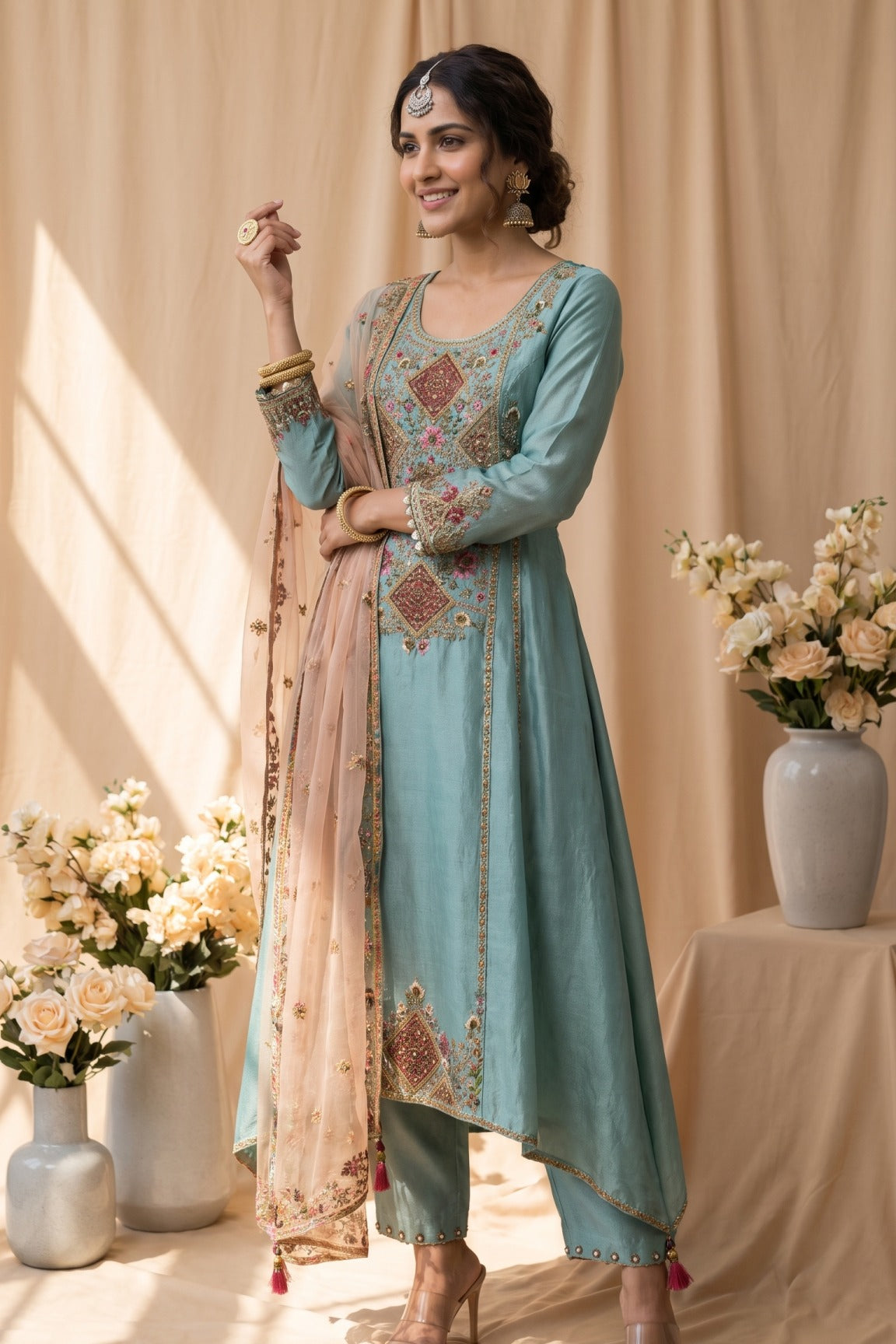 A light blue ethnic salwar kameez suit displayed on a Girl, featuring intricate multicolor floral embroidery on the chest, hem, and sleeves, along with a matching sheer dupatta draped over one shoulder. The background is a warm beige studio setting with draped curtains and decorative floral arrangements.