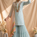  displayed on a girlagainst a draped beige fabric backdrop, with floral embroidery on the neckline, a matching flared skirt, and a sheer dupatta with pastel multicolor borders and gold trim. Decorative flower arrangements sit on both sides. blue kurta set 