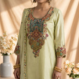A light yellow-green unstitched salwar kameez suit set displayed on a mannequin, featuring a heavily embroidered kameez top with a multicolor floral neckline, matching wide-leg trousers, and a sheer printed dupatta draped over the arm, set against a warm beige background with decorative floral arrangements.