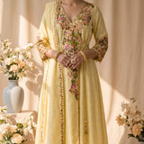 A light yellow traditional kurta set with colorful floral embroidery displayed on a mannequin. The set includes a long kurta with a V-neck, long sleeves, and a self-tie waist, paired with matching trousers.