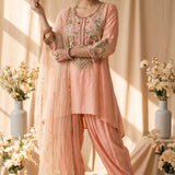 A peach-pink three-piece salwar kameez suit set displayed on a mannequin, featuring a kurta with intricate floral embroidery on the chest, matching draped pants, and a sheer dupatta, set against a warm beige backdrop with decorative floral arrangements.