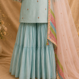 A light blue traditional outfit with a tiered flared skirt, embroidered top, and a sheer peach dupatta with a multicolored patterned border, displayed against a warm beige backdrop.
