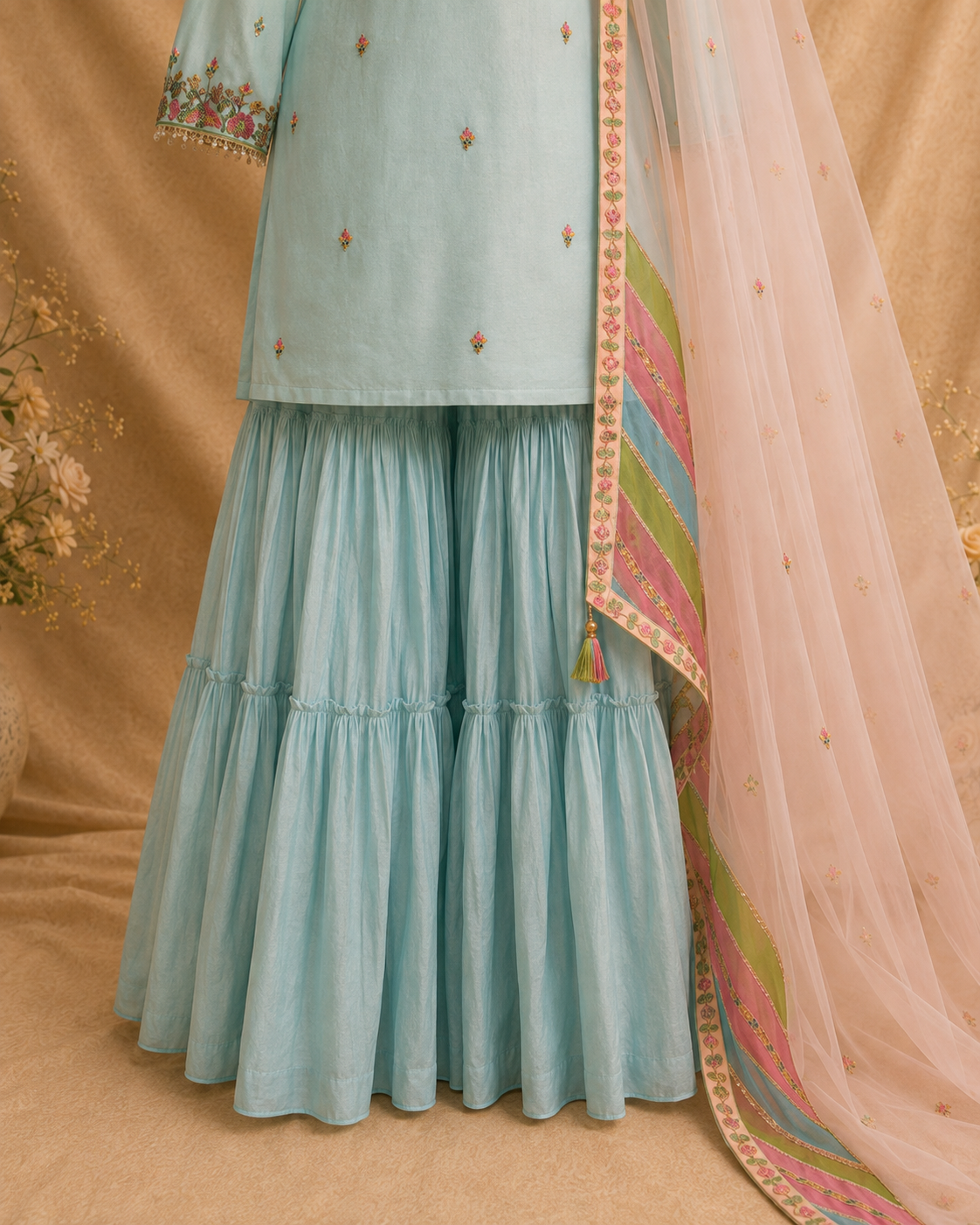 A light blue traditional outfit with a tiered flared skirt, embroidered top, and a sheer peach dupatta with a multicolored patterned border, displayed against a warm beige backdrop.