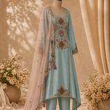 A light blue traditional outfit displayed on a mannequin, featuring ornate multicolor embroidery, matching pants, and a sheer peach dupatta with gold and floral detailing, set against a warm beige backdrop with draped fabric and flower arrangements.