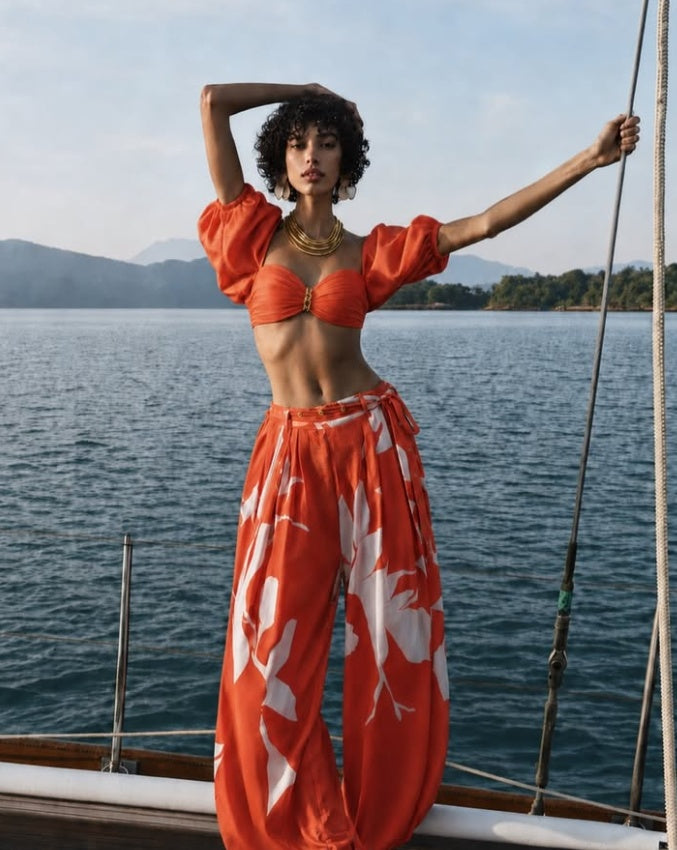 A woman stands on a boat wearing a bright orange two-piece outfit with puff sleeves and a matching top, paired with wide-leg pants featuring a white floral print. The background shows blue water and distant hills under a pale sky. resort wear mandira wirk 