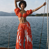 A woman stands on a boat wearing a bright orange two-piece outfit with puff sleeves and a matching top, paired with wide-leg pants featuring a white floral print. The background shows blue water and distant hills under a pale sky. resort wear mandira wirk 