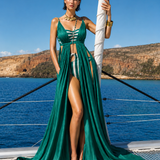 A model standing on a yacht wears a flowing emerald green resort gown with a deep neckline, front cutouts, and a striped swimsuit-style inner detail, styled with gold jewelry against a sea and coastal backdrop.