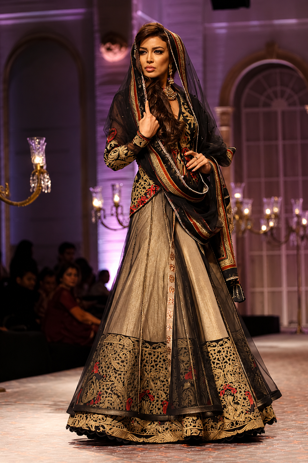 A model walks the runway wearing an ornate traditional South Asian outfit with a beige and black embroidered skirt, a heavily embellished black dupatta, and a gold-trimmed embroidered bodice. The background shows a fashion show venue with chandeliers and seated audience members.