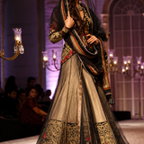 A model walks the runway wearing an ornate traditional South Asian outfit with a beige and black embroidered skirt, a heavily embellished black dupatta, and a gold-trimmed embroidered bodice. The background shows a fashion show venue with chandeliers and seated audience members.