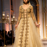A model walks the runway wearing a long-sleeved beige and gold embellished Anarkali gown with a sheer tulle skirt, set against a grand indoor venue with candelabras.