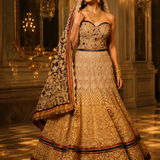 A woman models an ornate traditional Indian lehenga choli set in a grand, warmly lit palace hall. The outfit is heavily embellished with gold embroidery and sequins, featuring a strapless bodice, a full flared skirt, and a matching dupatta draped over her shoulder.