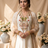 A traditional white salwar kameez suit displayed on a mannequin, featuring intricate multicolor floral embroidery on the chest and dupatta, set against a warm beige textured backdrop with decorative floral arrangements.