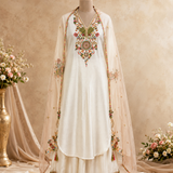 A traditional white salwar kameez suit displayed on a mannequin, featuring intricate multicolor floral embroidery on the chest and dupatta, set against a warm beige textured backdrop with decorative floral arrangements.
