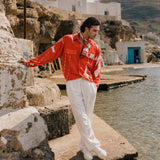 A man stands on a stone pier by the water wearing a bright red long-sleeve button-up shirt with white graphic panels and white trousers, with a coastal village and rocky cliffs in the background. Resort wear man 