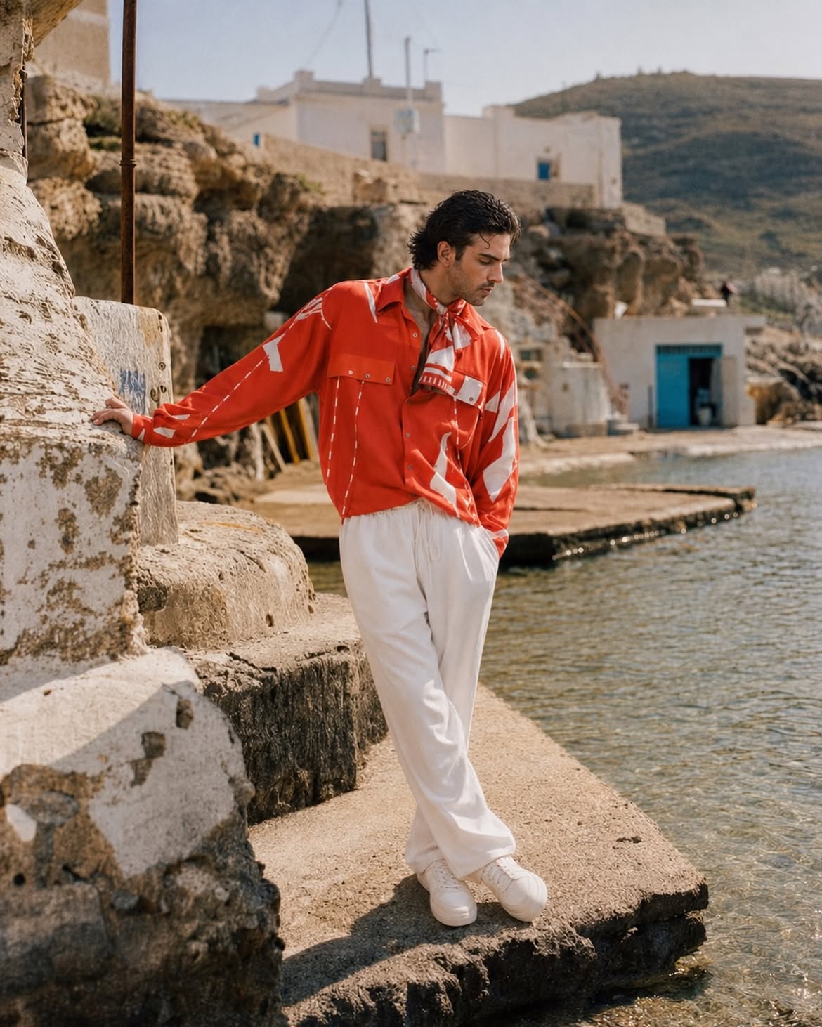 A man stands on a stone pier by the water wearing a bright red long-sleeve button-up shirt with white graphic panels and white trousers, with a coastal village and rocky cliffs in the background. Resort wear man 