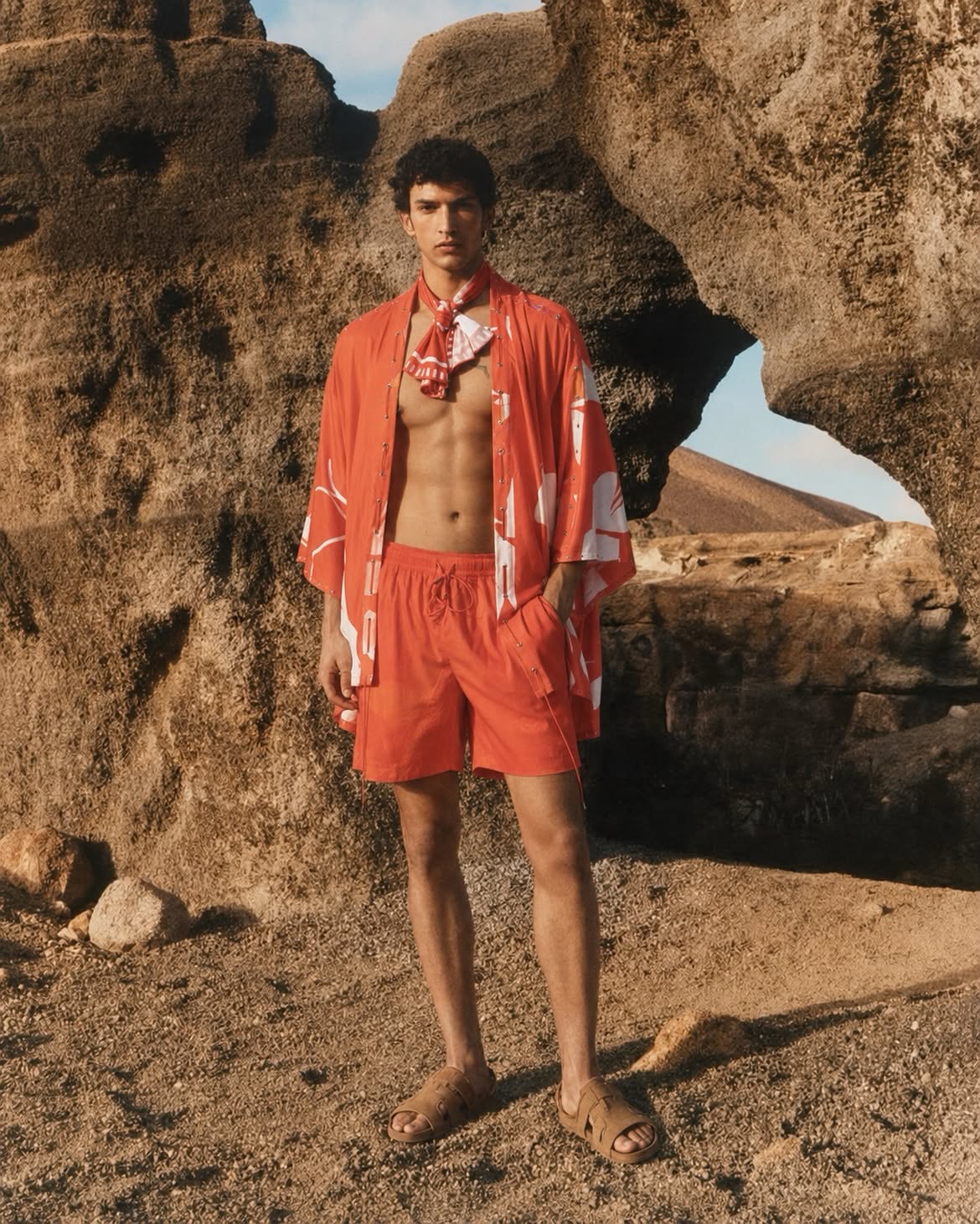 A male model stands outdoors in a desert rock formation wearing a matching orange-red swim set with a white abstract pattern, including a short-sleeve open-front top, shorts, and a neck scarf, along with tan sandals. resort wear 