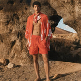 A male model stands outdoors in a desert rock formation wearing a matching orange-red swim set with a white abstract pattern, including a short-sleeve open-front top, shorts, and a neck scarf, along with tan sandals. resort wear 