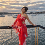 A woman wearing a red and white patterned one-shoulder maxi dress stands on a waterfront balcony at sunset, with boats and water in the background.