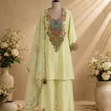 A light yellow-green unstitched salwar kameez suit set displayed on a mannequin, featuring a heavily embroidered kameez top with a multicolor floral neckline, matching wide-leg trousers, and a sheer printed dupatta draped over the arm, set against a warm beige background with decorative floral arrangements.