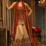 A woman models a traditional South Asian outfit consisting of a beige flared skirt with colorful floral embroidery, a matching embroidered top, and a long red and gold embroidered scarf, set against an elegant indoor background with a chandelier.