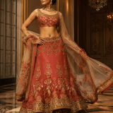 A woman modeling a traditional Indian lehenga choli set in a grand, ornate hall. The outfit is a coral pink with extensive gold embroidery, featuring a matching sheer dupatta draped over her arm.