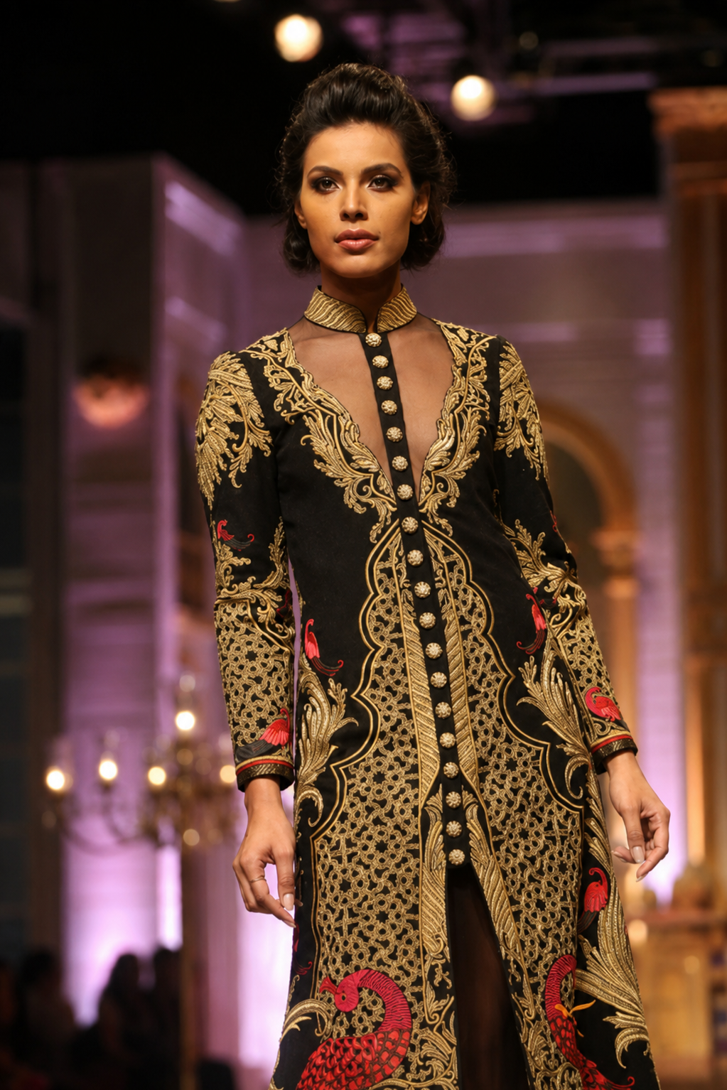 A model walks the runway wearing a long-sleeved black and gold patterned tunic dress with red accents, featuring a sheer neckline and a buttoned front.