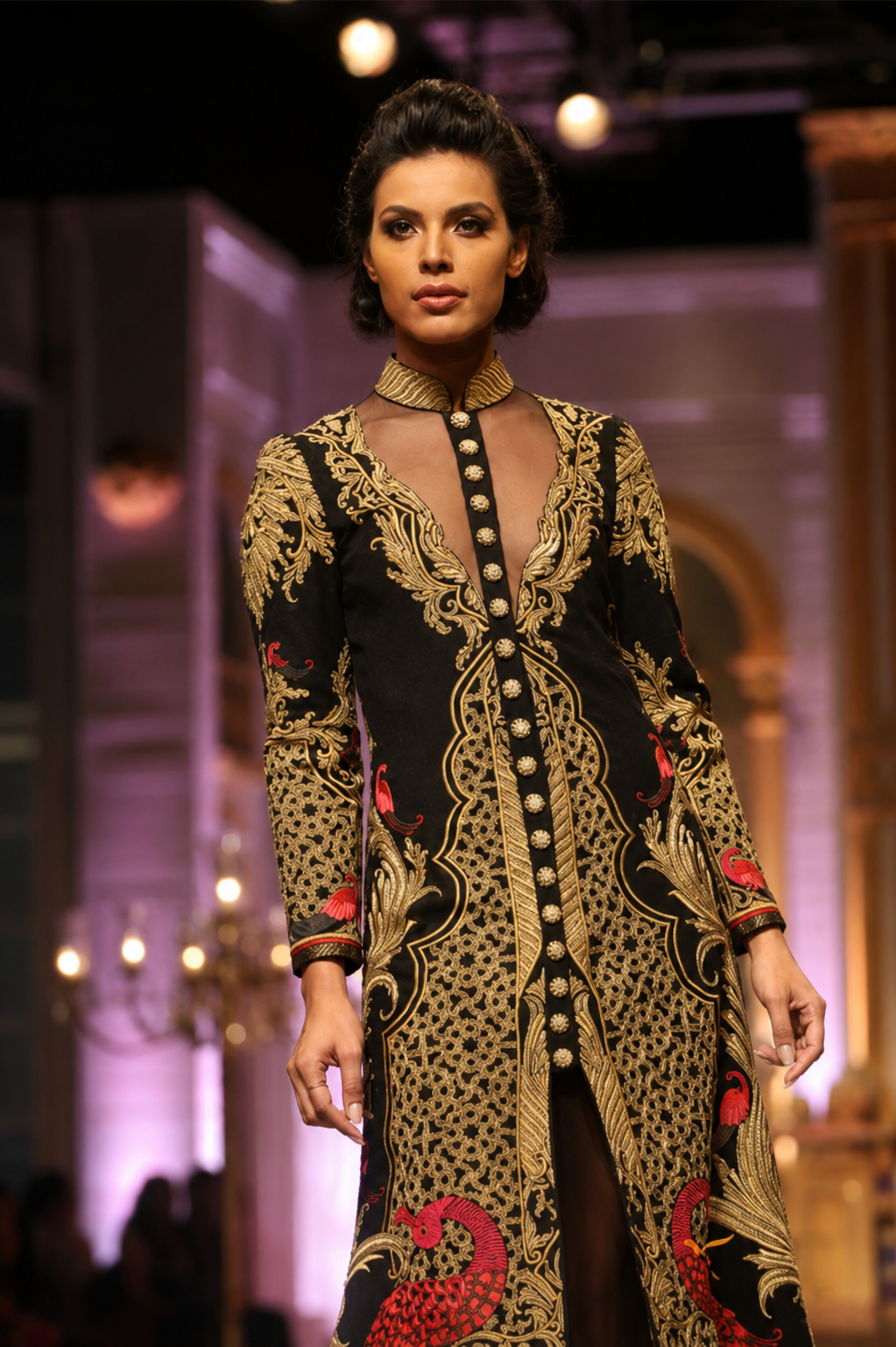 A model walks the runway wearing a long-sleeved black and gold patterned tunic dress with red accents, featuring a sheer neckline and a buttoned front.
