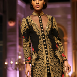 A model walks the runway wearing a long-sleeved black and gold patterned tunic dress with red accents, featuring a sheer neckline and a buttoned front.
