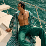 Male model wearing emerald green trousers with a flowing green-and-white striped resort overlay by Mandira Wirk, seated aboard a luxury yacht.