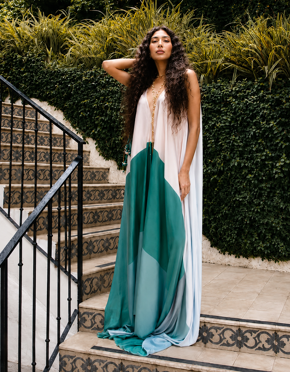 Model wearing a flowing white, teal and aqua colour-block resort kaftan by Mandira Wirk, styled with gold jewellery on patterned outdoor steps.
