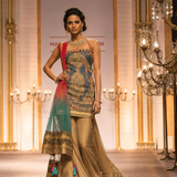 A model walks a fashion runway wearing a traditional Indian outfit with a heavily embroidered blue and gold sleeveless top, beige flared pants, and a red-to-teal gradient dupatta.