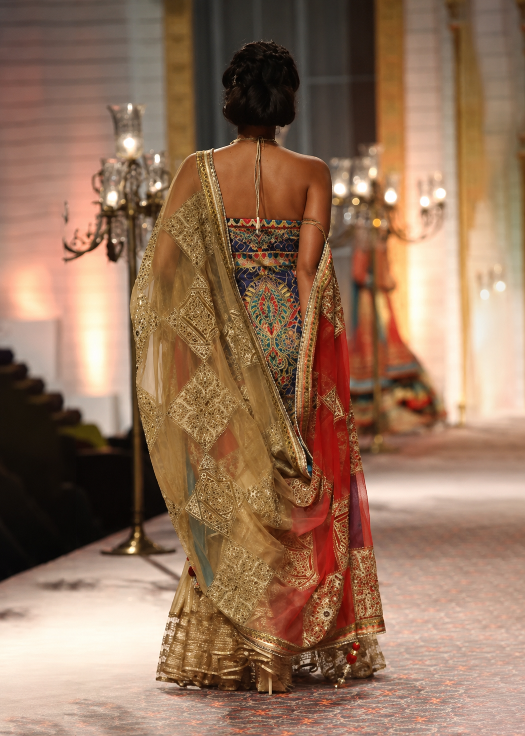A model viewed from behind walking on a runway wearing an ornate traditional Indian lehenga choli. The garment features a highly detailed multicolor embroidered bodice with a sheer gold embroidered dupatta draped over her shoulders.