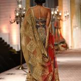 A model viewed from behind walking on a runway wearing an ornate traditional Indian lehenga choli. The garment features a highly detailed multicolor embroidered bodice with a sheer gold embroidered dupatta draped over her shoulders.